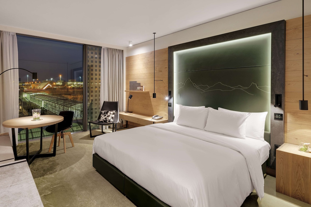 Habitación de hotel moderna con una cama grande, un escritorio, una silla, una pequeña mesa con un florero y vistas a un paisaje urbano nocturno.