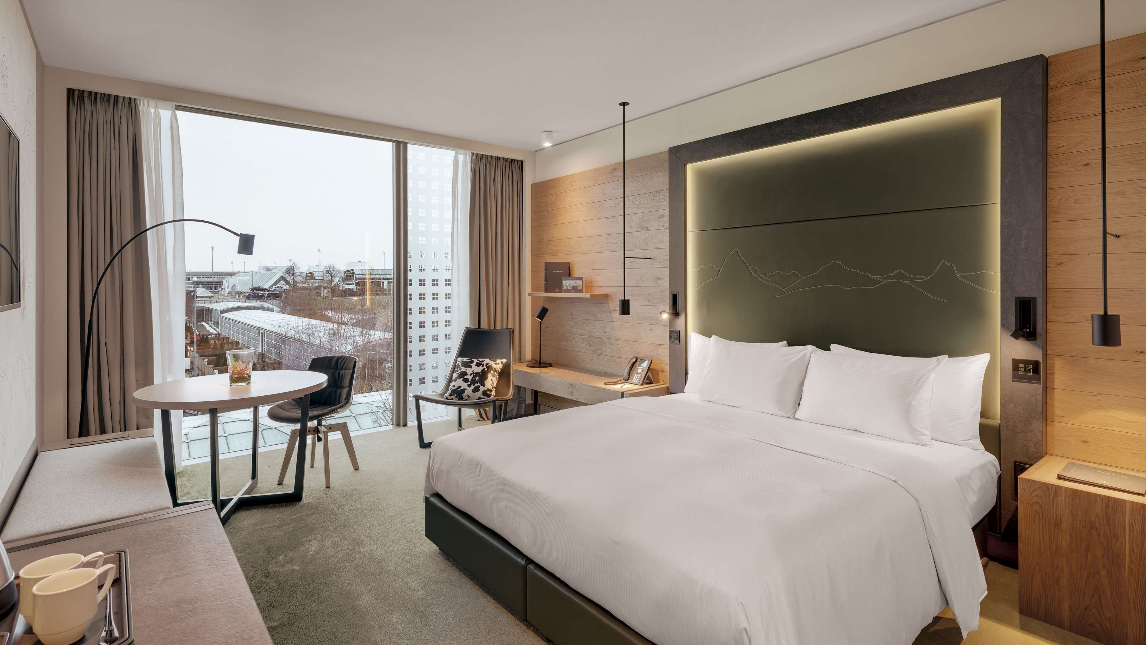Habitación de hotel moderna con una cama grande, un escritorio, una silla y vistas a la ciudad a través de amplias ventanas.
