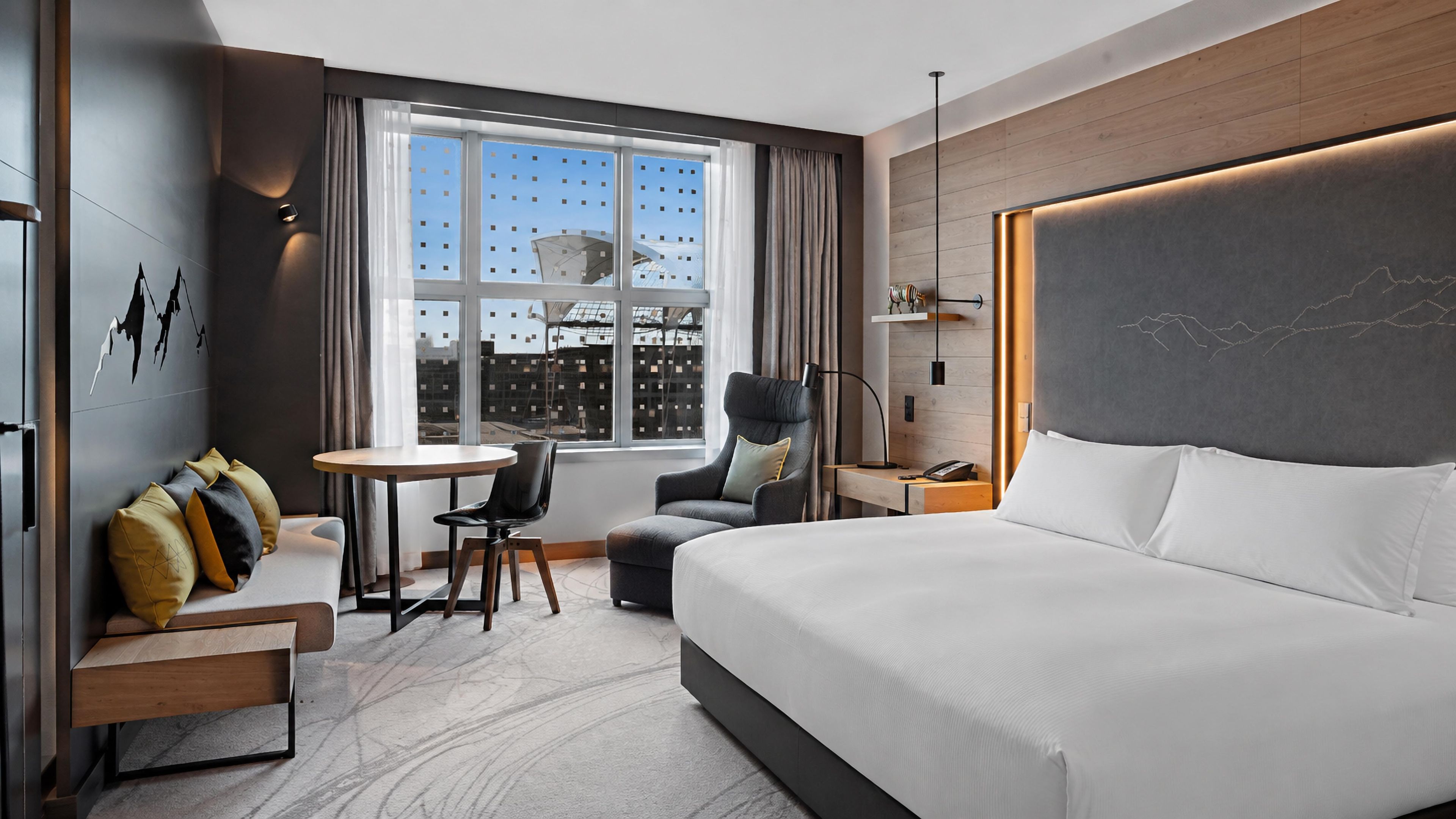 Ein modernes Hotelzimmer mit einem großen Bett, einem Schreibtisch, einem Stuhl und Blick auf die Stadtlandschaft durch das Fenster.