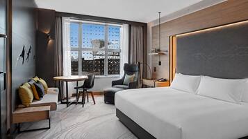 Ein modernes Hotelzimmer mit einem großen Bett, einem Schreibtisch, einem Stuhl und Blick auf die Stadtlandschaft durch das Fenster.
