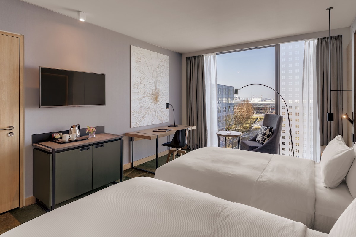 A hotel room with a bed, desk, and chair, a TV, and a large window with a city view.