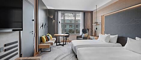 A hotel room with a large bed, a smaller bed, a desk, a chair, a sofa, a TV, and a window with a city view.