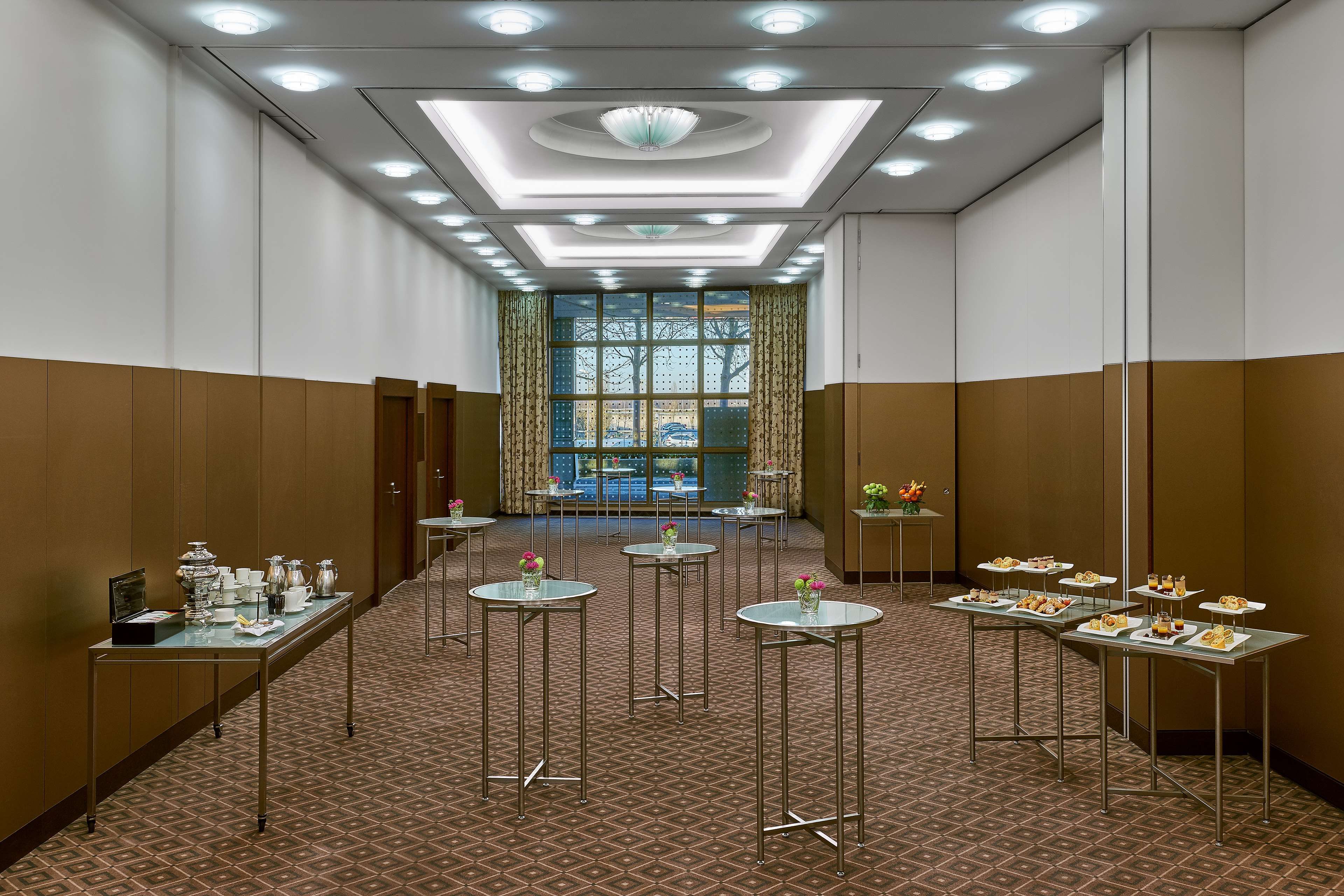 Ein Hotelgang mit Teppichmuster, runden Tischen mit Glasplatten und Blick ins Freie durch große Fenster.