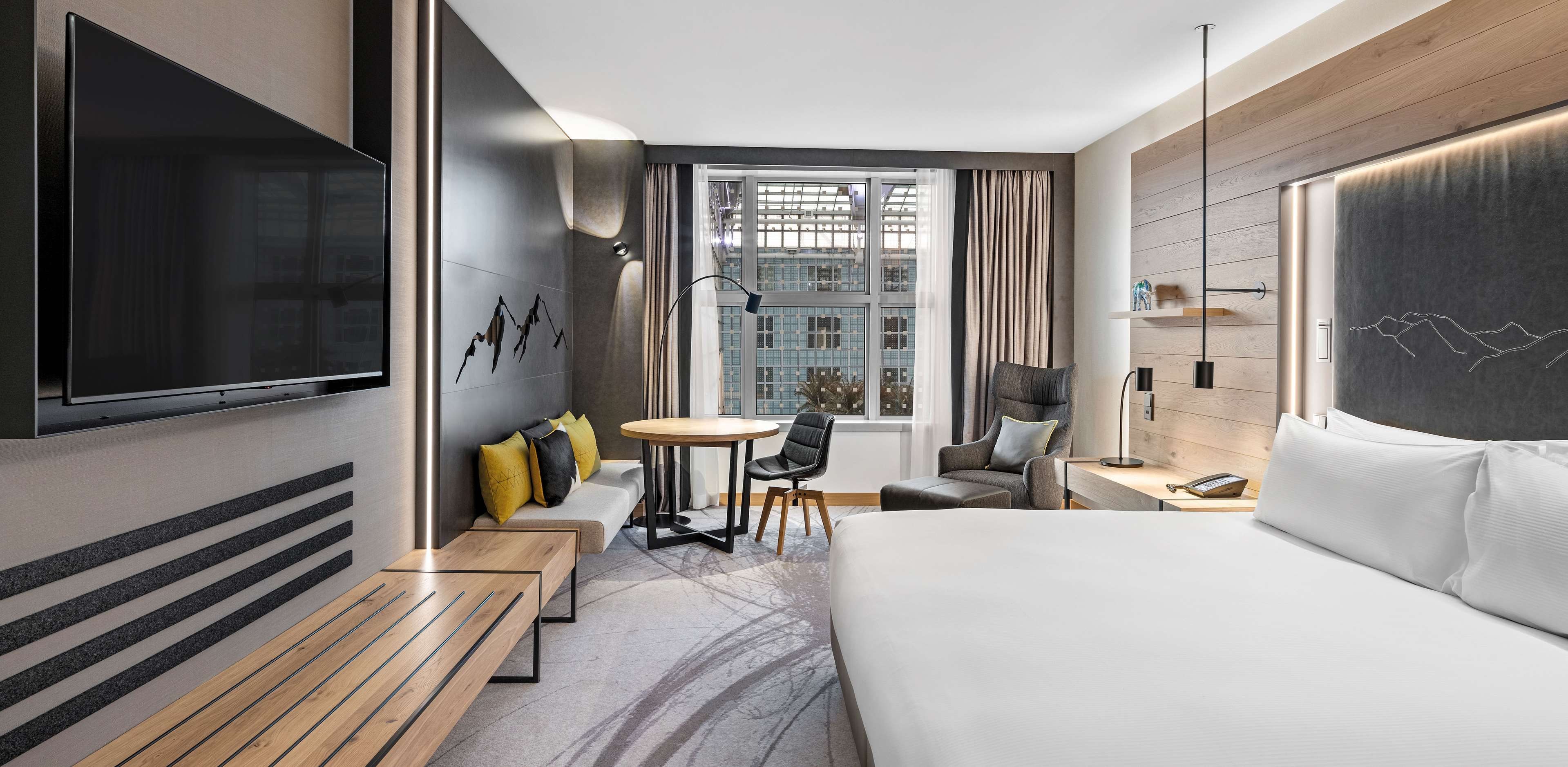 Habitación de hotel moderna con una cama grande, televisor de pantalla plana, mesa de comedor y vistas a la ciudad.