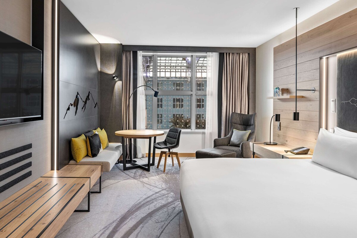 Una habitación de hotel moderna con una cama grande, un televisor de pantalla plana en la pared, una pequeña mesa redonda con sillas, un sofá con cojines y una ventana con cortinas.