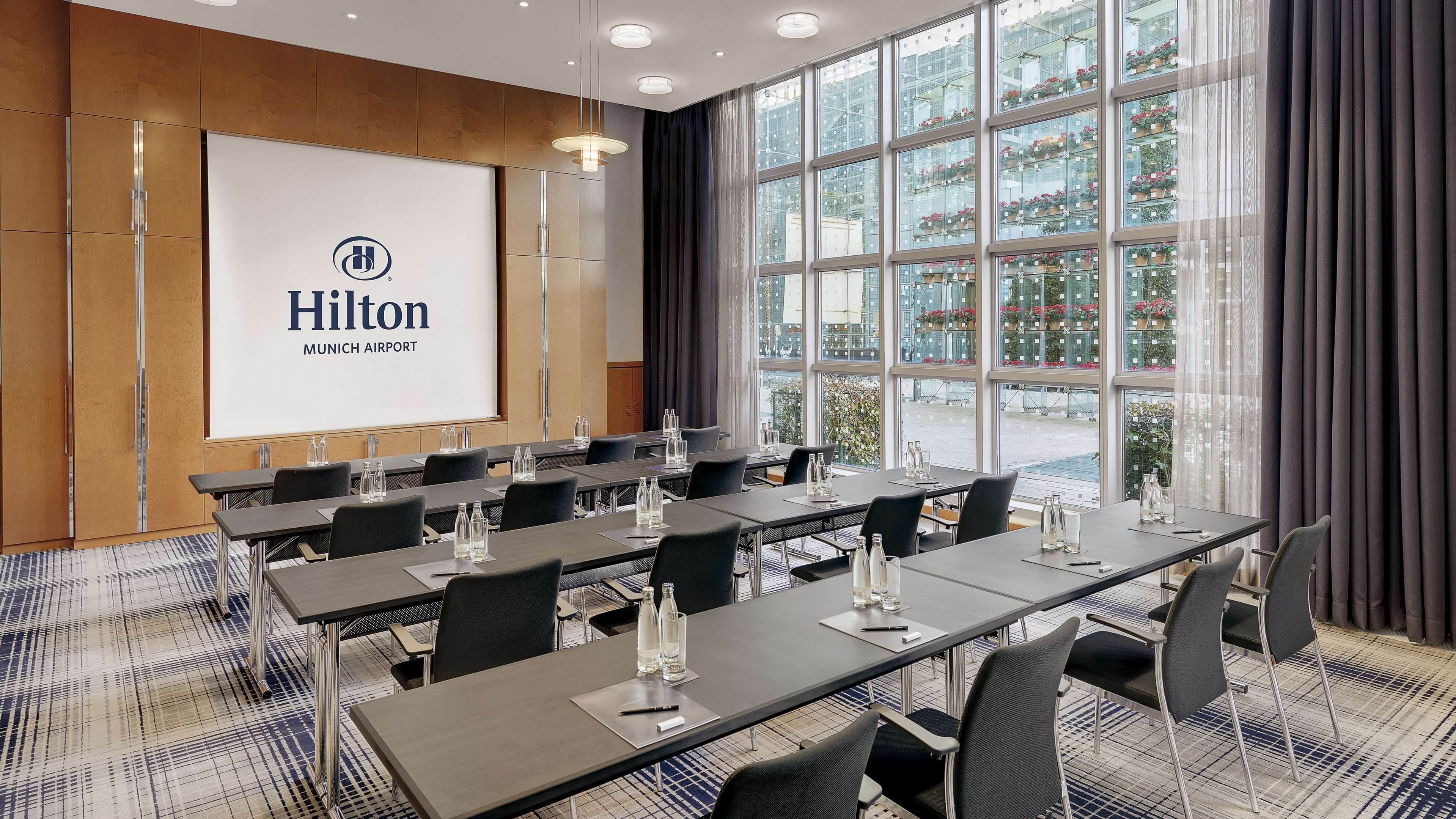 Un salón de conferencias en el Hilton del Aeropuerto de Múnich con varias mesas, sillas y una pantalla grande mostrando el logotipo de Hilton.