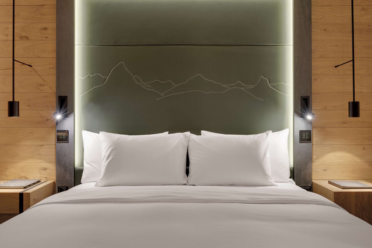Una habitación de hotel moderna con una cama grande, ropa de cama blanca y un cabecero con diseño de paisaje montañoso. Hay dos mesitas de noche con lámparas y un panel de control en el cabecero.