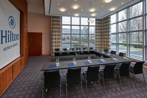 Un salón de conferencias con una larga mesa, sillas y una pantalla proyectora que muestra 'Hilton Munich Airport'.