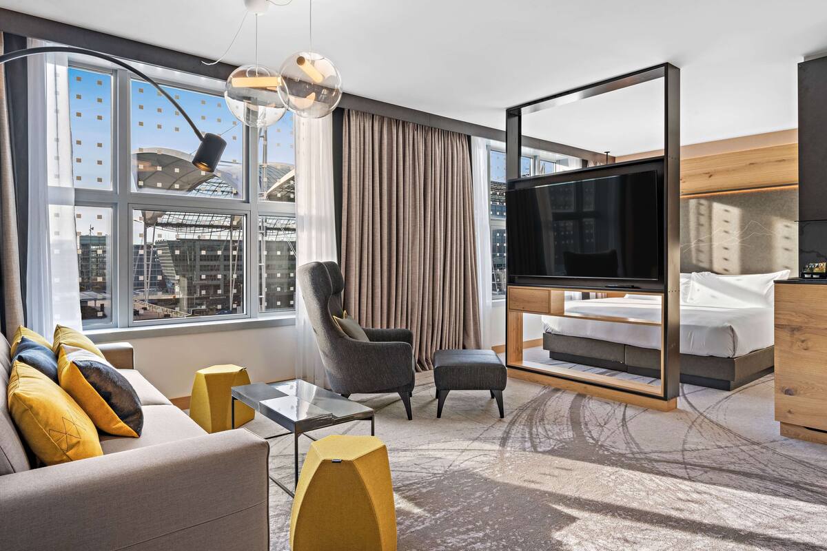A modern hotel room with a large window, a sofa with cushions, a chair, a TV, a bed, and a wooden headboard.