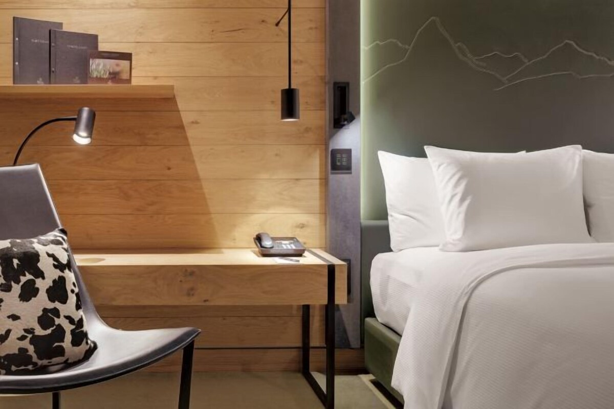 A hotel room with a wooden desk, a chair with a cow-patterned cushion, a bed with white bedding, and a wall with a mountain line design.