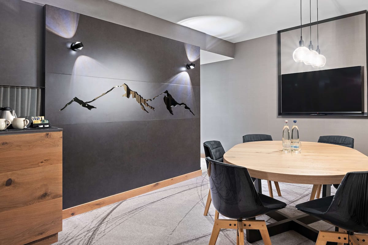 A conference room with a round table, black chairs, a wooden cabinet, a wall-mounted TV, and a ceiling light fixture.