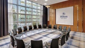 Un salón de conferencias con una gran mesa ovalada, múltiples sillas, una pantalla de proyector con el logotipo de Hilton y ventanas amplias con vistas a edificios y árboles.
