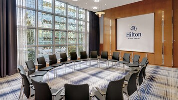 Un salón de conferencias con una gran mesa ovalada, múltiples sillas, una pantalla de proyector con el logotipo de Hilton y ventanas amplias con vistas a edificios y árboles.