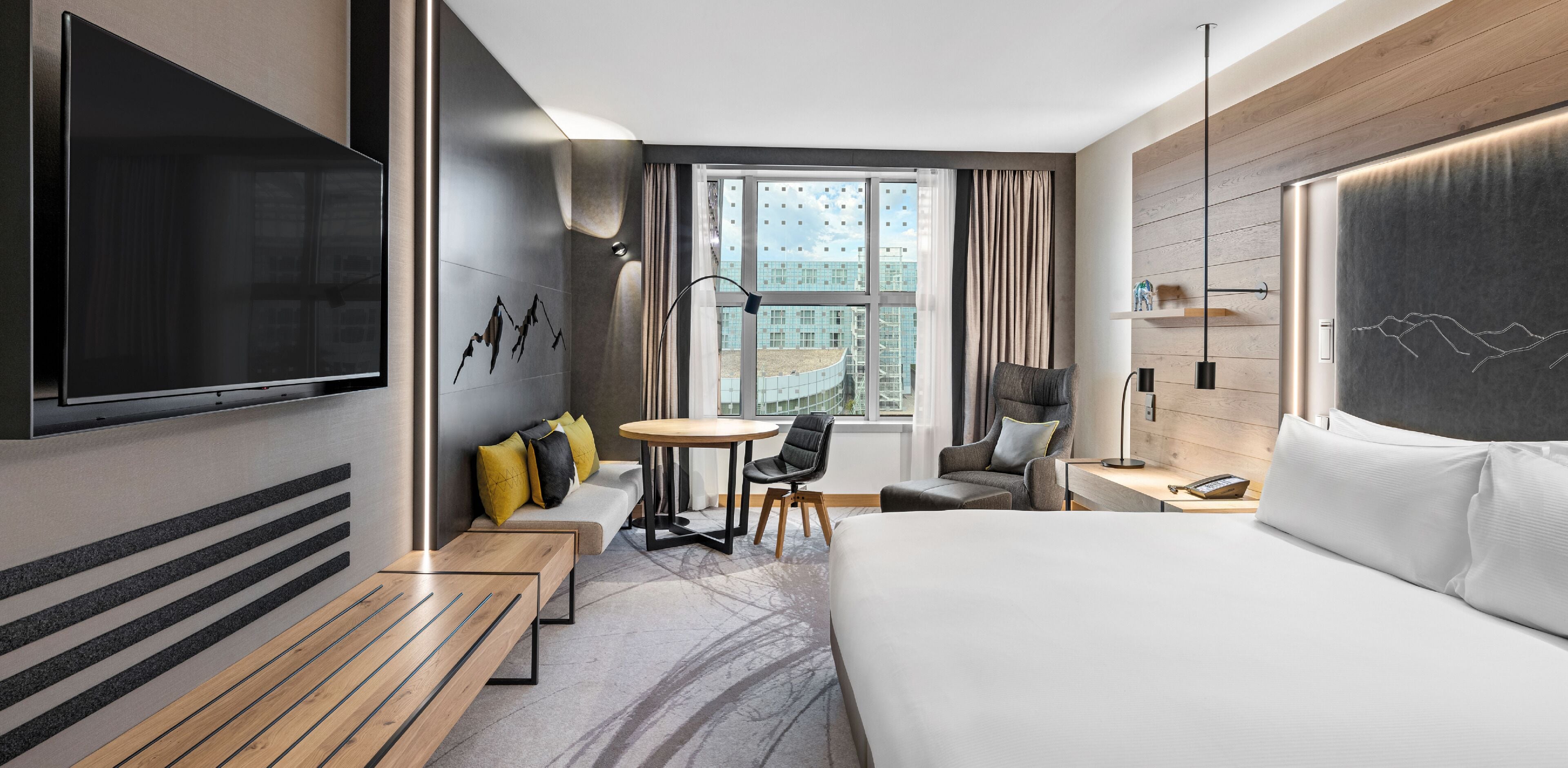 A hotel room with a bed, a desk, a chair, a small table, and a TV mounted on the wall.