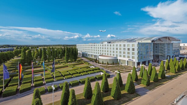 Un hotel moderno con fachada de vidrio, rodeado de un jardín cuidado y varias banderas.