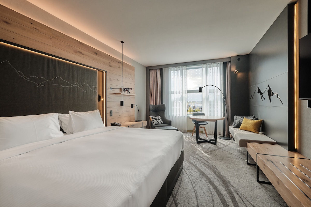 A modern hotel room with a large bed, a desk, a sofa, and a view of the city.