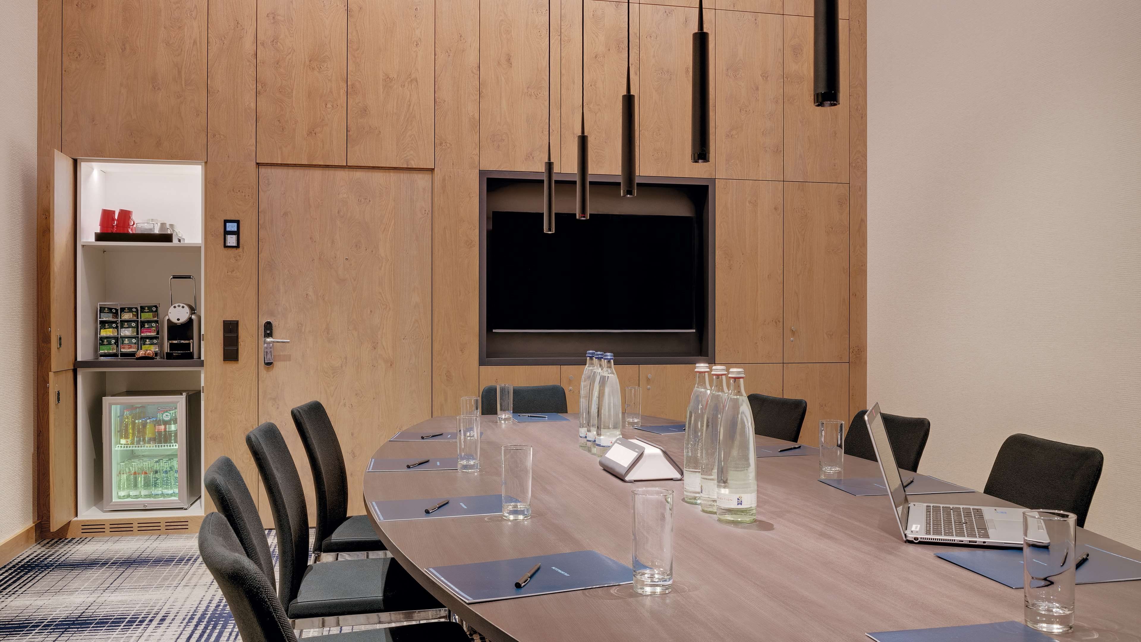 Un salón de conferencias con una gran mesa, sillas, una pantalla de proyector y un refrigerador con bebidas.