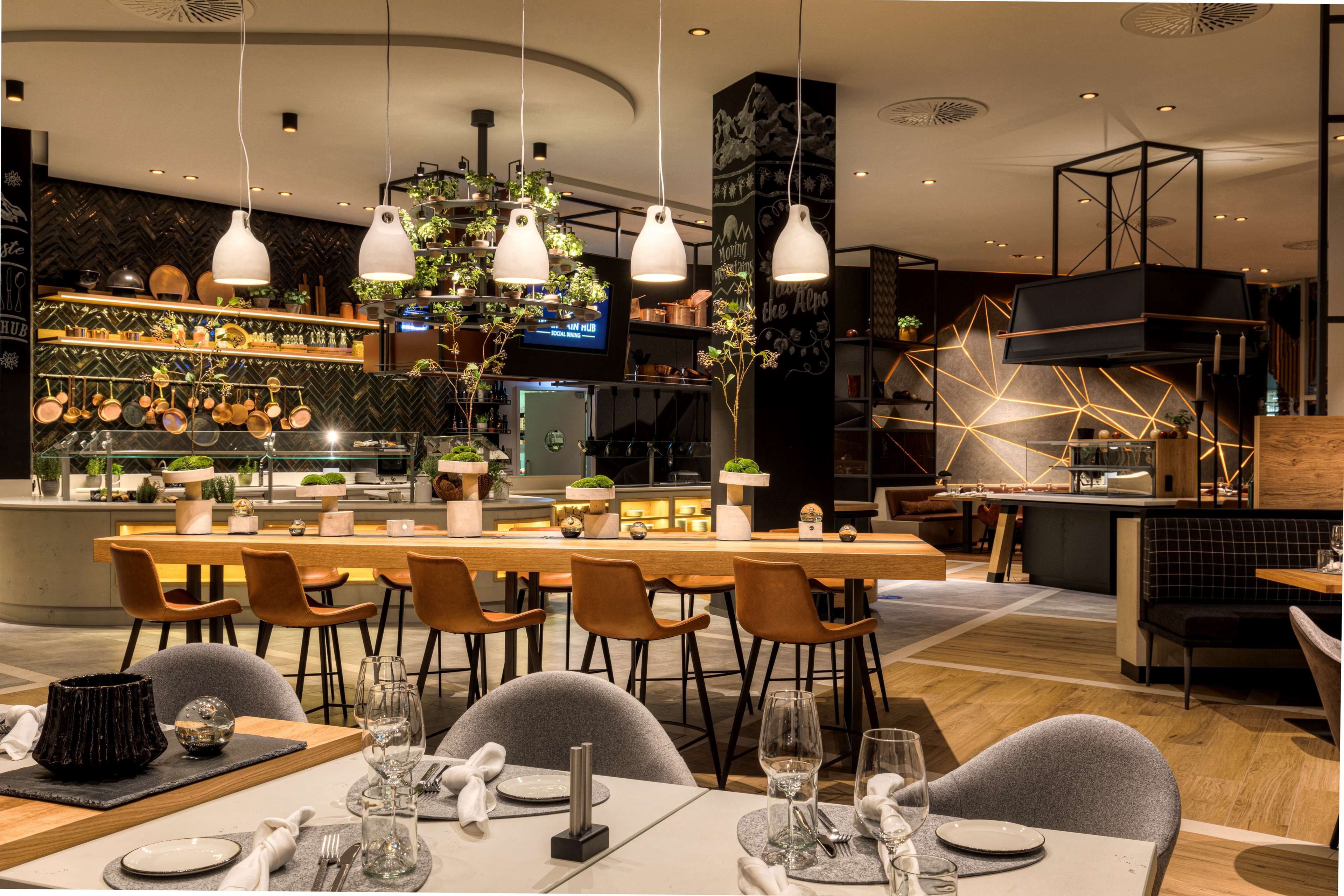 A modern restaurant interior with a central dining area, kitchen in the background, and a bar area with a geometric light installation.
