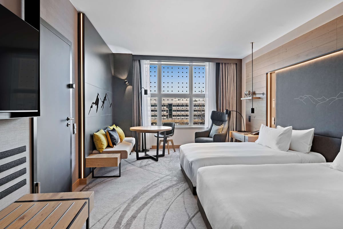 A modern hotel room with a large bed, a smaller bed, a sofa, a desk, and a TV. There is a window with a city view and a wall with a mountain design.