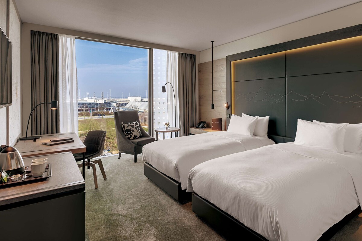 Habitación de hotel con dos camas, un escritorio con lámpara, una silla y vistas a la ciudad.