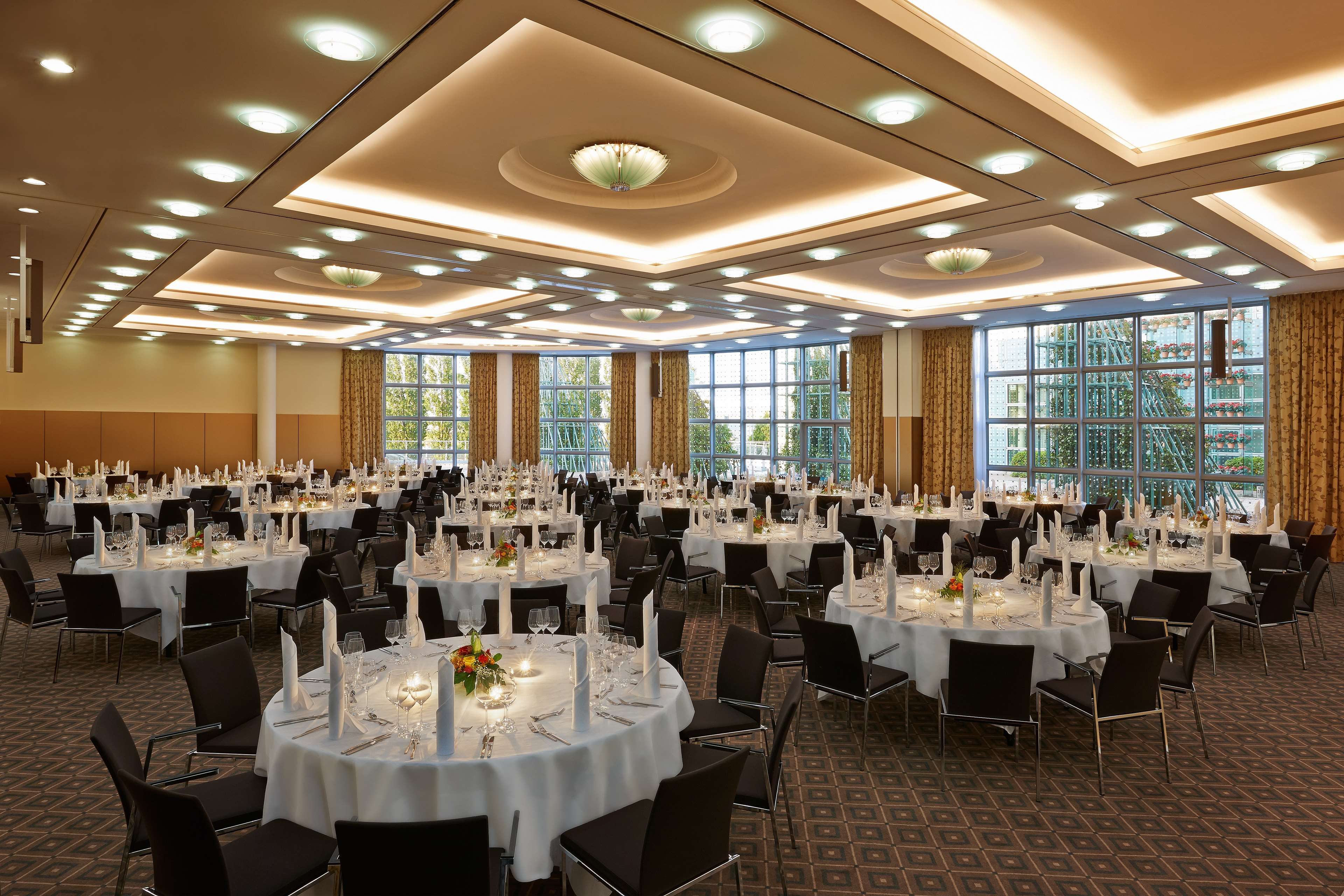 A banquet hall with round tables set for dining, large windows, and a high ceiling with recessed lighting.