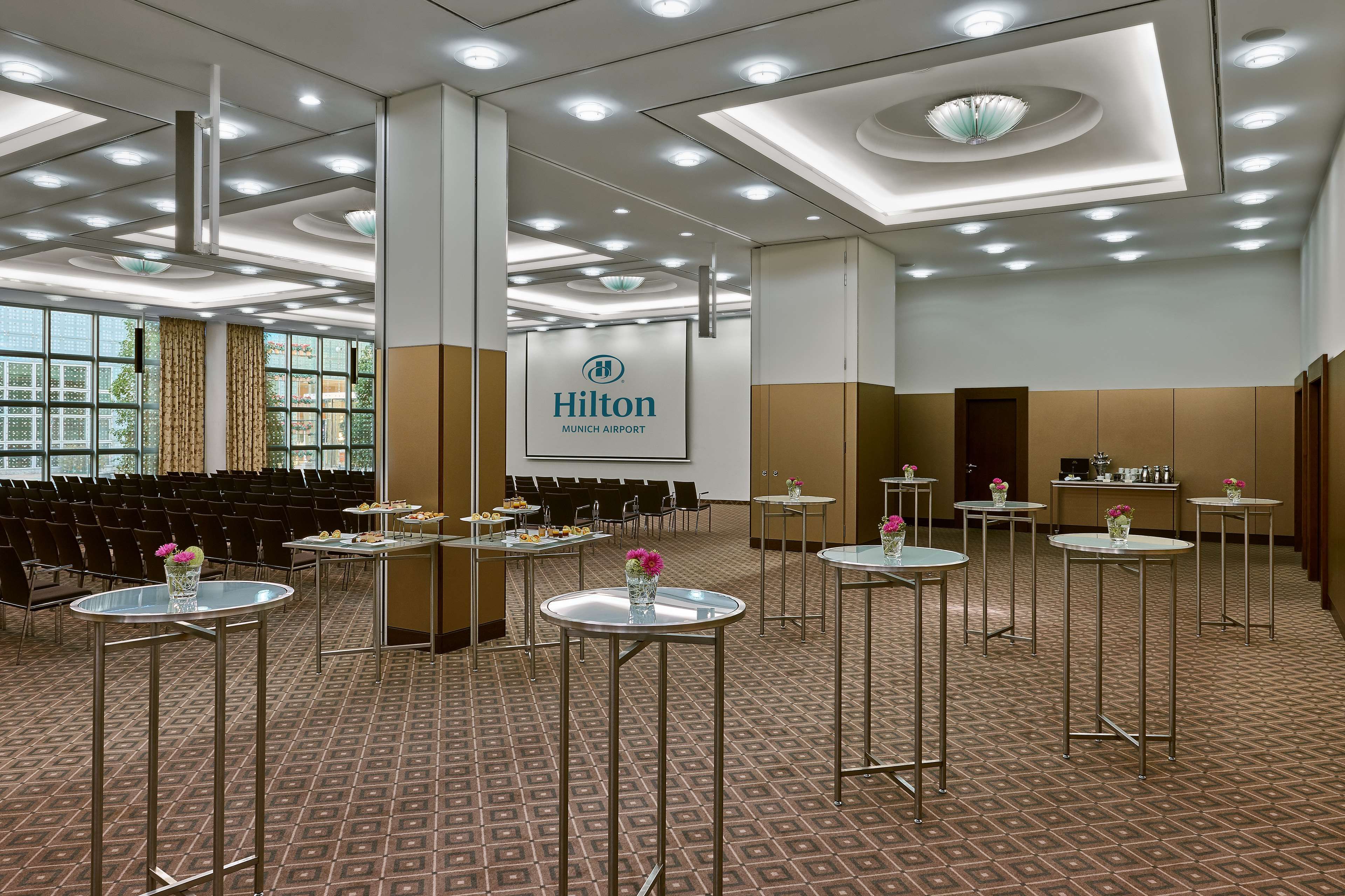 Uma sala de conferências de hotel com uma tela grande, várias mesas com arranjos florais e uma área de estar com fileiras de cadeiras.