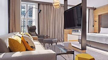 Habitación de hotel moderna con una cama grande, un sofá con almohadones decorativos, un televisor, una mesita y vistas a la ciudad a través de amplios ventanales.