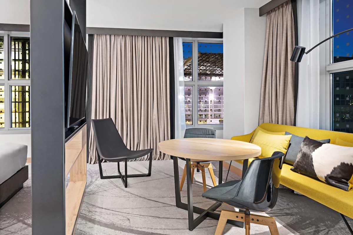 A modern hotel room with a yellow sofa, a round wooden table, and two black chairs.