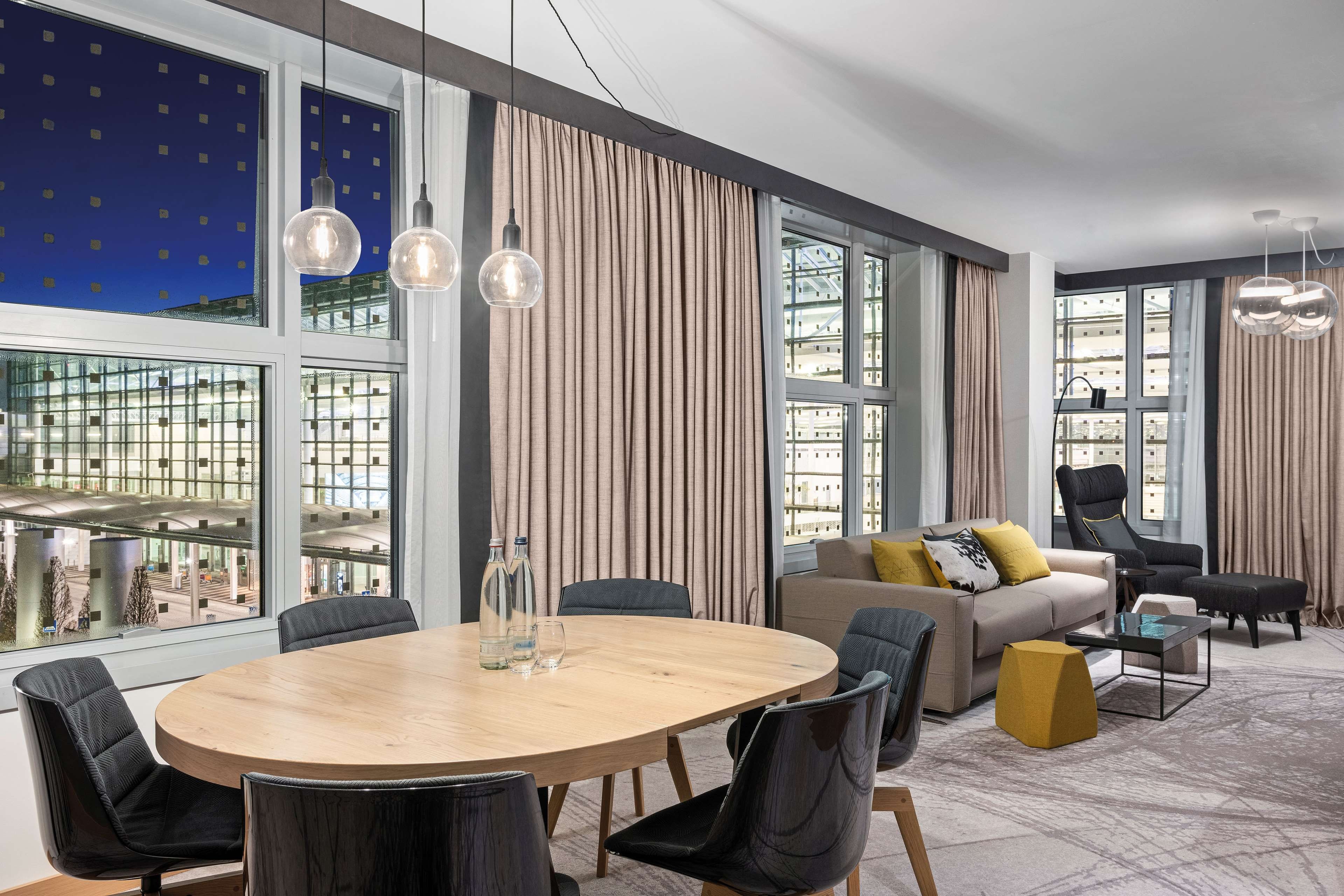 A modern hotel room with a round dining table, multiple chairs, a sofa with cushions, a black armchair, and a glass coffee table. Large windows offer a view of the cityscape.