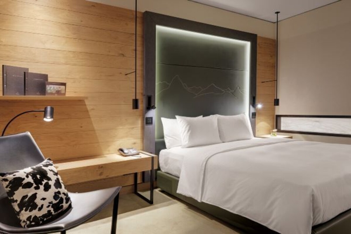 A modern hotel room with a wooden wall, a bed with white bedding, a desk with a chair, and a nightstand.