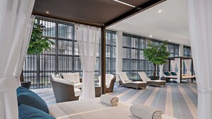 A modern hotel room with a large bed, a seating area with lounge chairs, and a view through large windows.