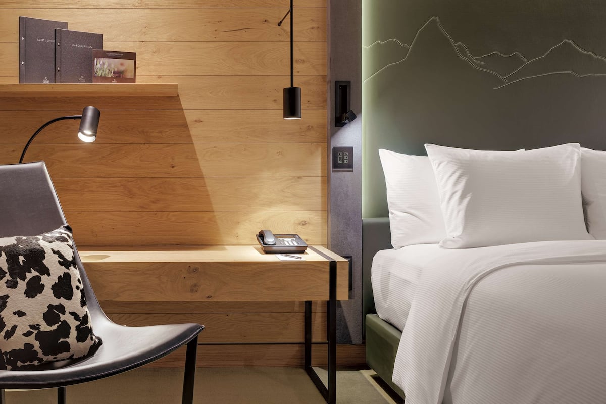 A modern hotel room with a wooden desk, a chair with a cow print cushion, a bed with white linens, and a wall with a mountain design.