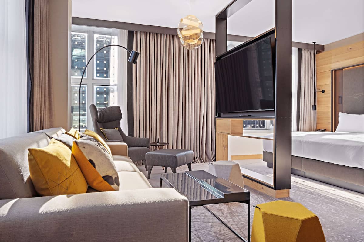 A modern hotel room with a sofa, armchair, and ottoman, a flat-screen TV, a bed, and a city view.