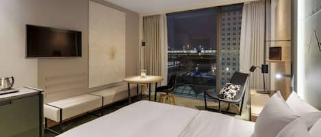 Habitació d'hotel amb un llit gran, un escriptori amb una làmpada, una cadira amb un coixí, una taula petita amb una làmpada i una televisió penjada a la paret.