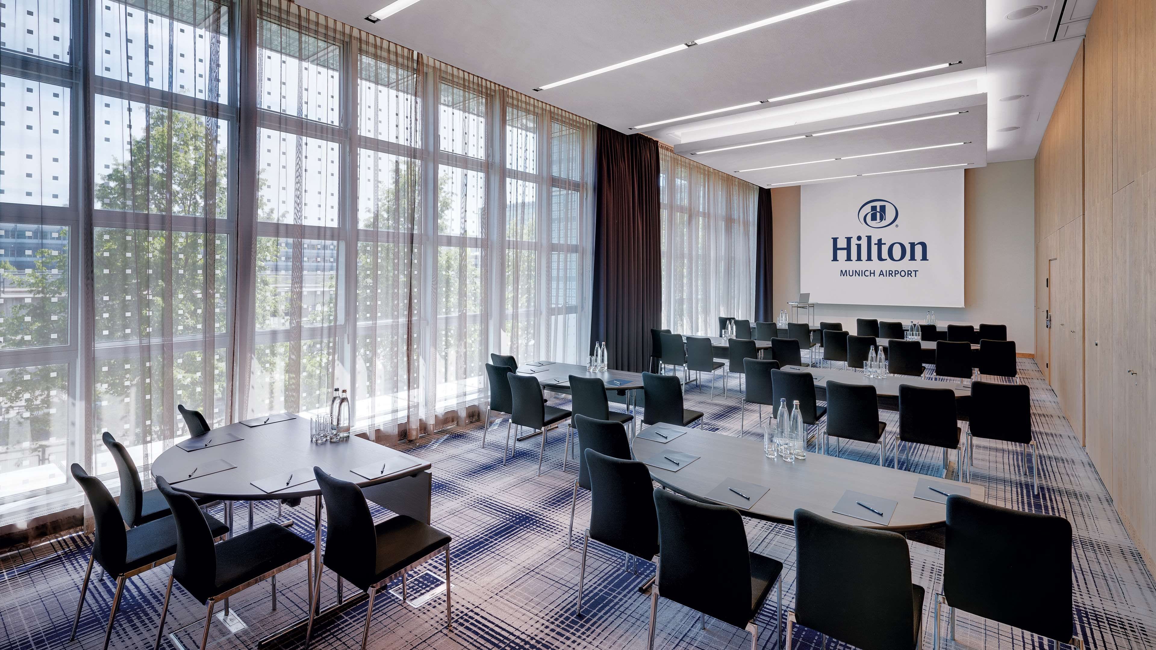 A conference room with tables and chairs, large windows with curtains, and a screen displaying 'Hilton Munich Airport'.