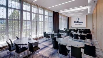 A conference room with tables and chairs, large windows with curtains, and a screen displaying 'Hilton Munich Airport'.