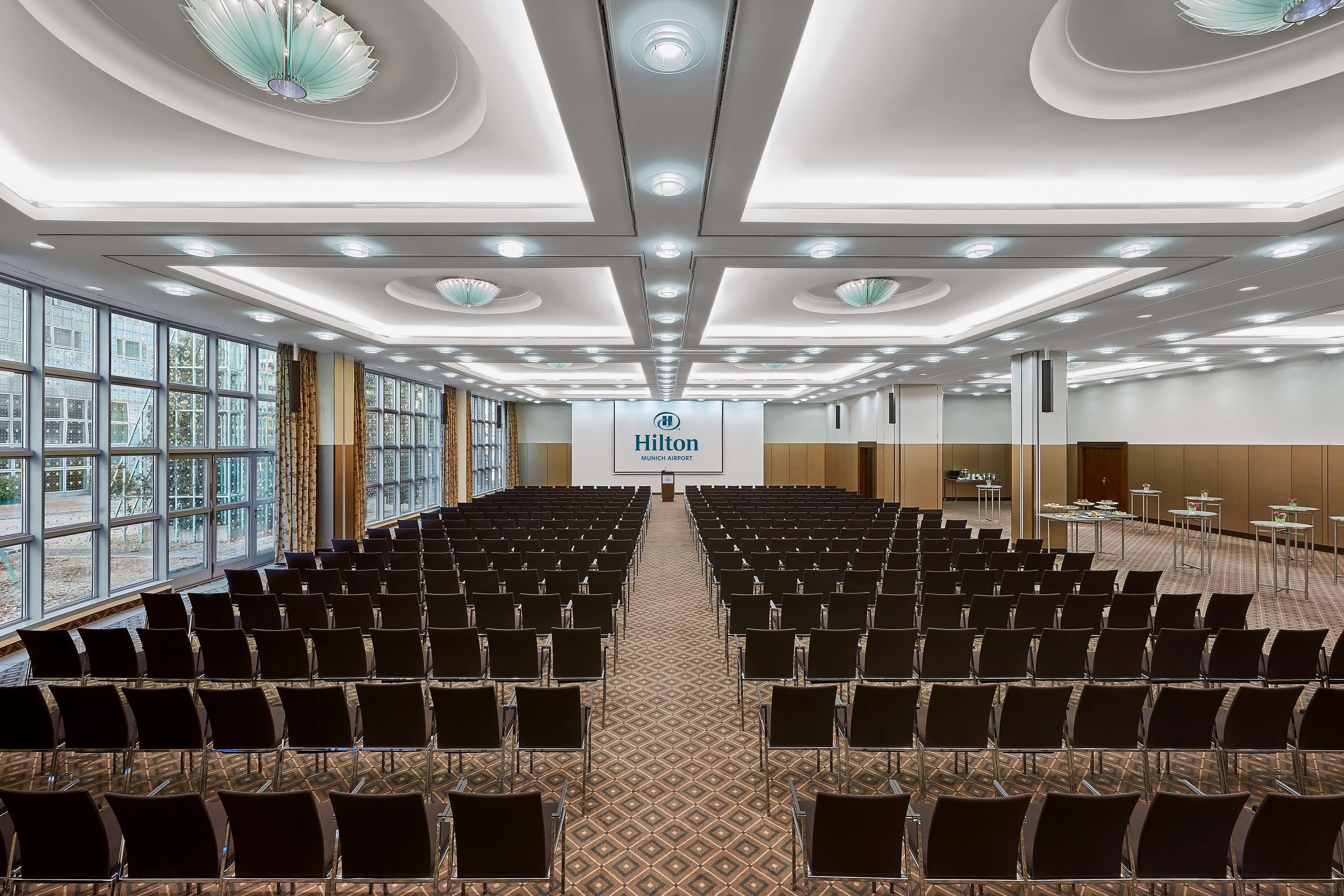 A large conference room with rows of chairs, a stage with a screen displaying the Hilton logo, and large windows providing natural light.