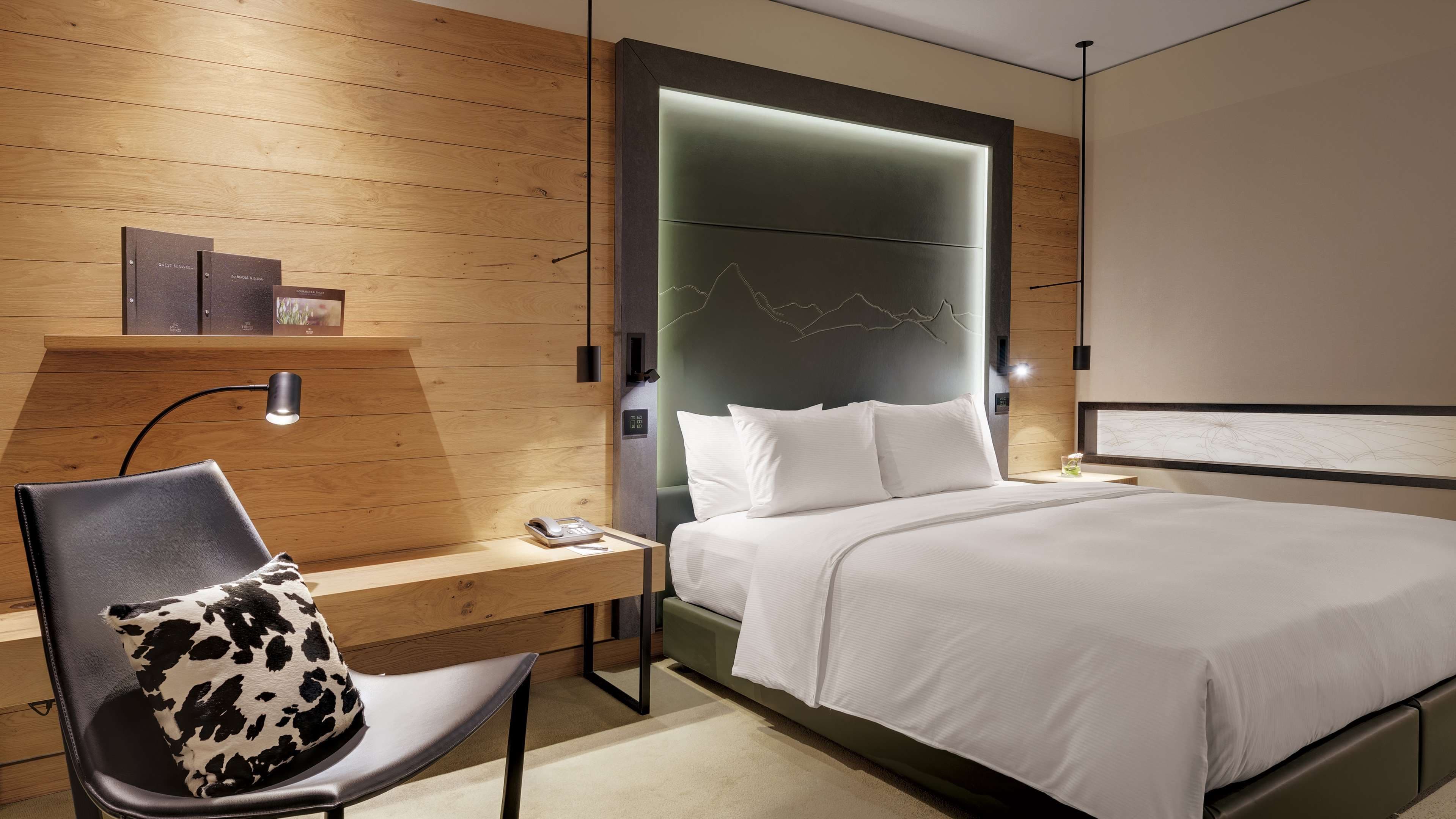 Ein modernes Hotelzimmer mit einer Wandpaneele aus Holz, einem Bett mit weißer Bettwäsche, einem Schreibtisch mit Lampe und einem Sessel mit Kissen mit Kuhmuster.