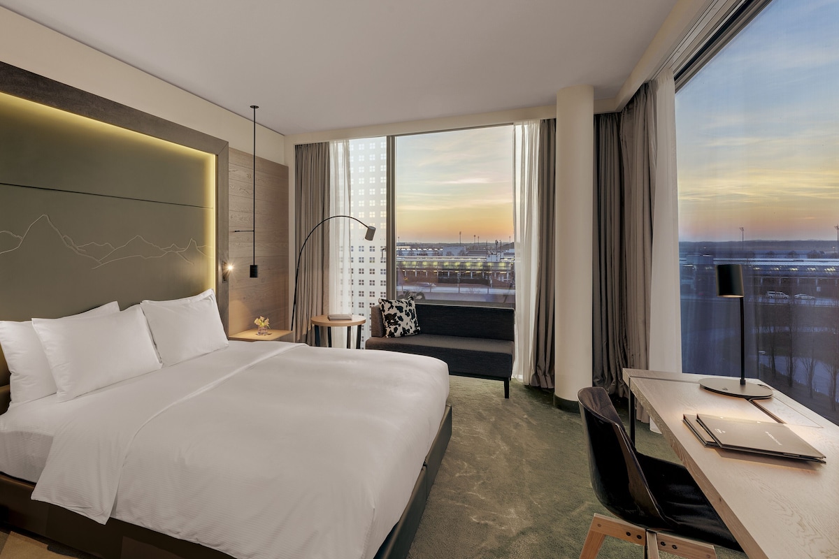 A modern hotel room with a large bed, a desk, and a view of the cityscape through large windows.