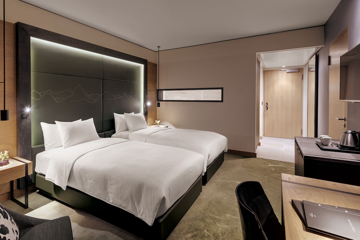 Ett modernt hotellrum med två sängar, ett skrivbord, en stol och en stor spegel.