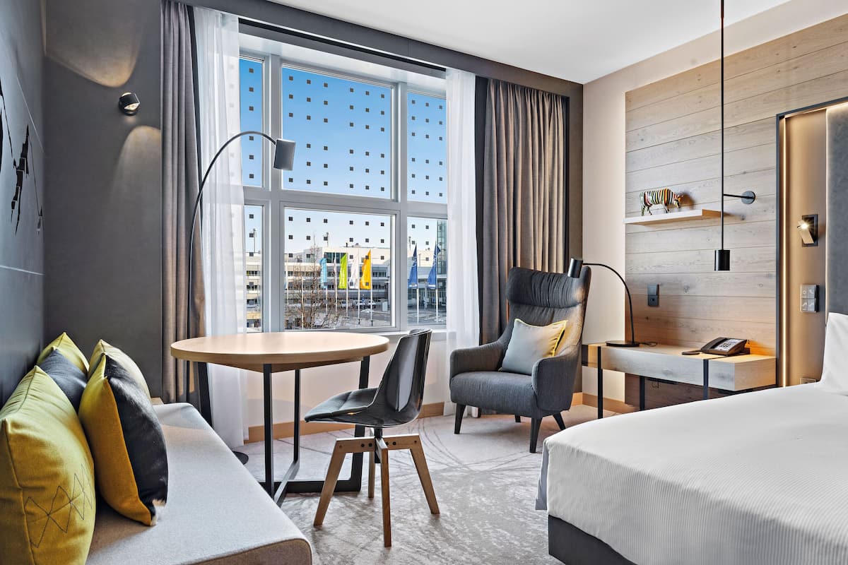 A modern hotel room with a large bed, a desk with a chair, a sofa with cushions, a large window with a view of buildings, and a wall-mounted shelf with decorative items.