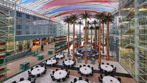 A spacious atrium with multiple dining tables and chairs, palm trees, and a central fountain.