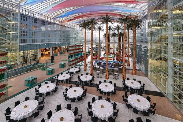 A spacious atrium with multiple dining tables and chairs, palm trees, and a central fountain. - Hilton Munich Airport (Oberding)
