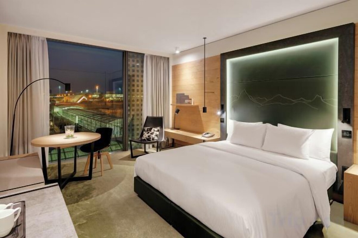 A hotel room with a large bed, a desk, a chair, a small table, and a view of a cityscape.