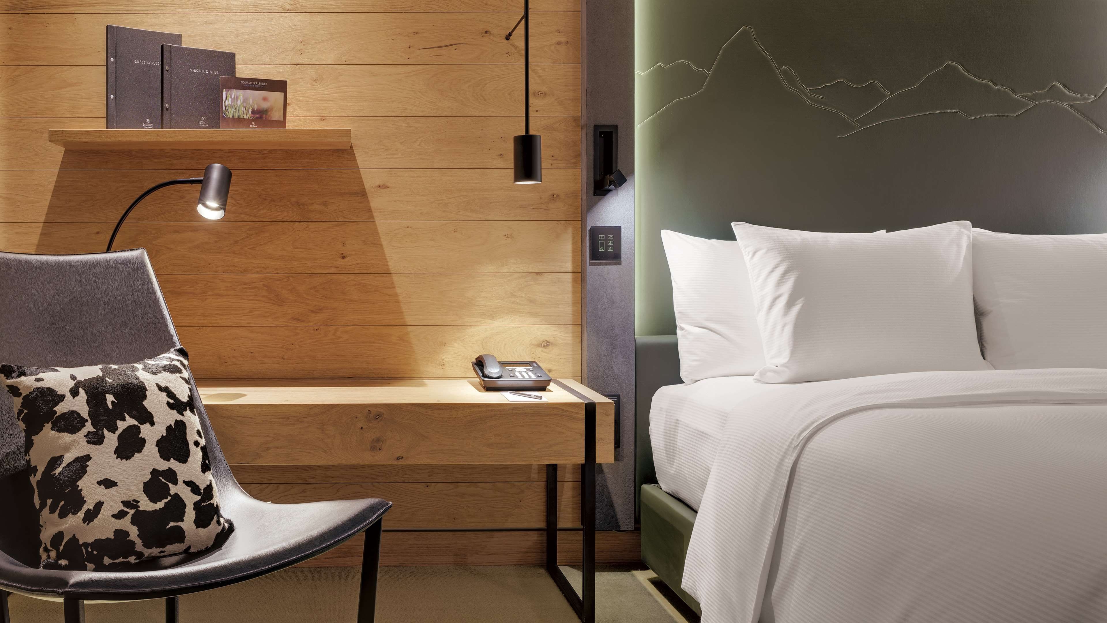 A hotel room with a wooden desk, a chair with a black and white patterned cushion, a bed with white bedding, and a wall-mounted phone.