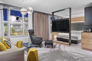 A modern living room with a sofa, armchair, ottoman, and a TV on a wooden stand. - Hilton Munich Airport (Oberding)