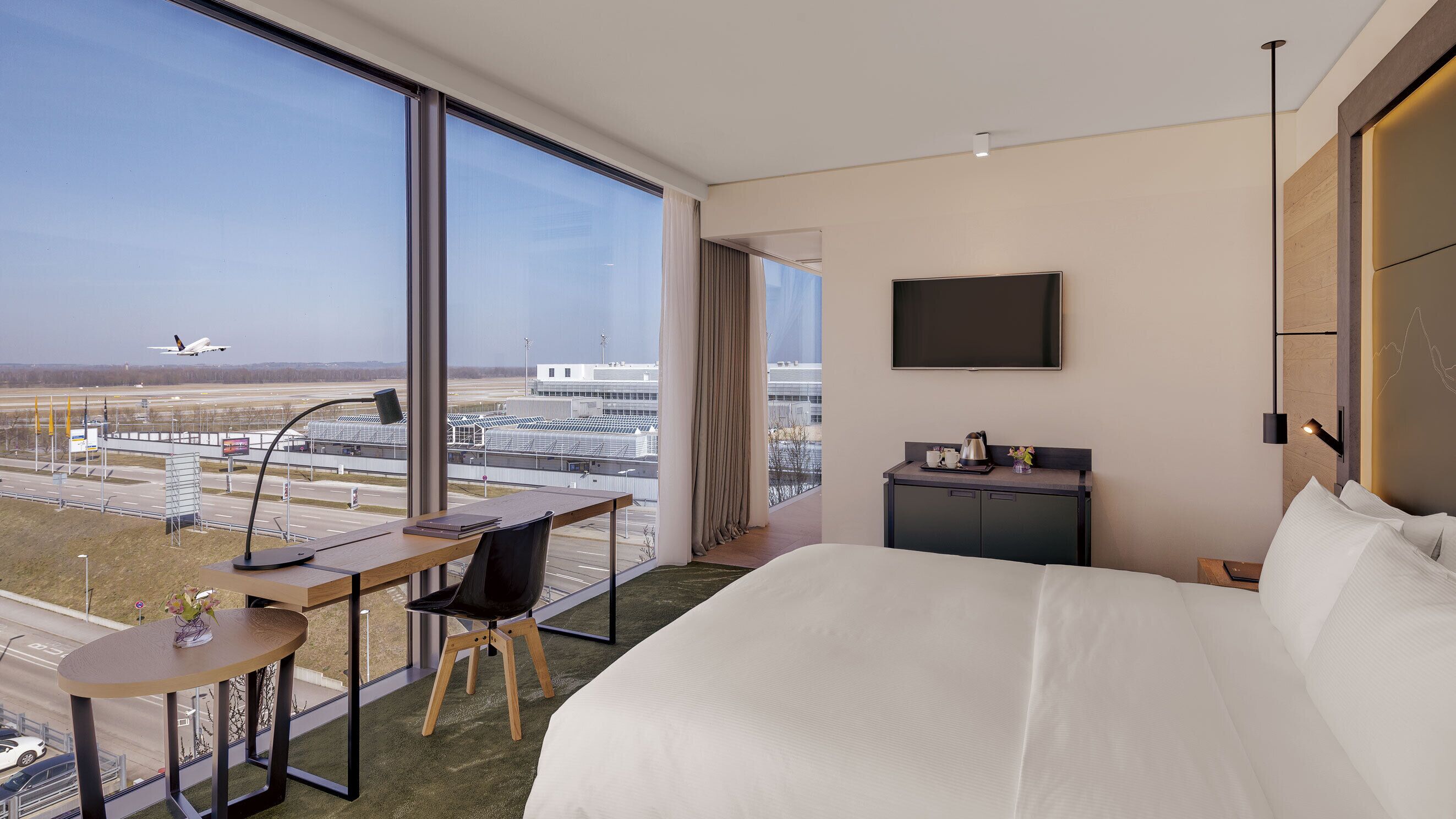 Een hotelkamer met een groot bed, een bureau met stoel, een televisie en uitzicht op een vliegveld.