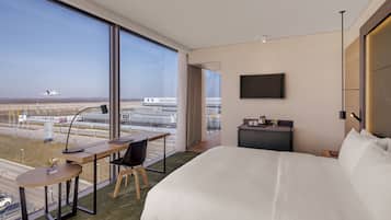 Een hotelkamer met een groot bed, een bureau met stoel, een televisie en uitzicht op een vliegveld.