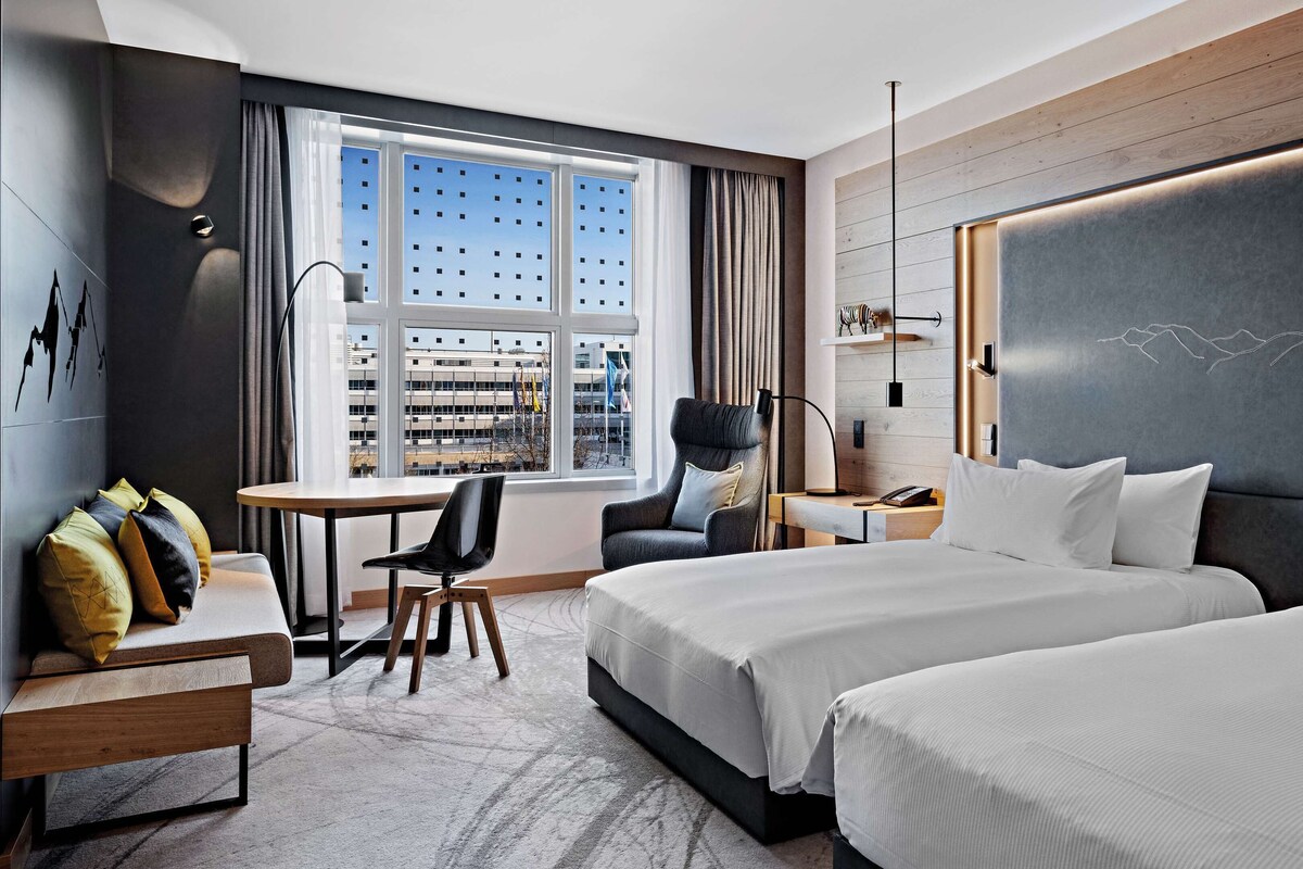 Habitación de hotel con dos camas, un escritorio, una silla y un amplio ventanal con vista a la ciudad.