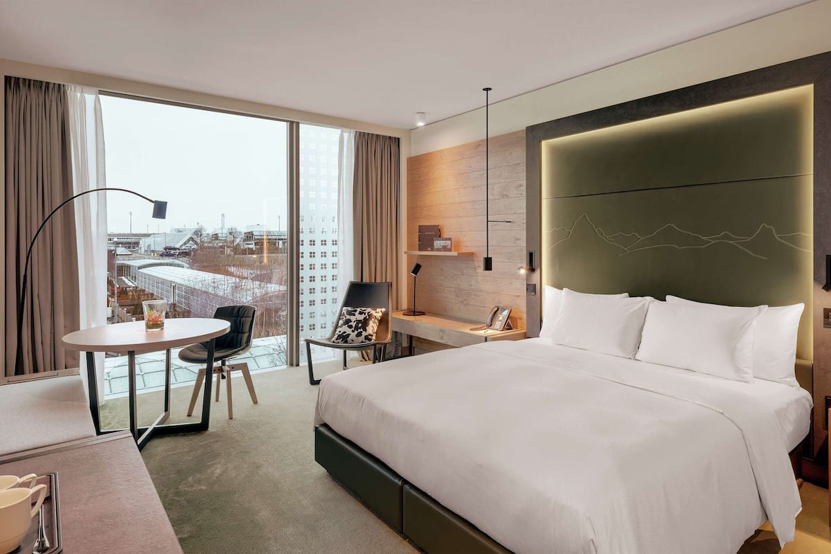 A modern hotel room with a large bed, a desk, a chair, and a view of the city through large windows.
